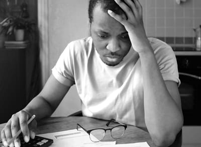 一个黑人坐在餐桌旁，双手抱着头，看着账单和文件