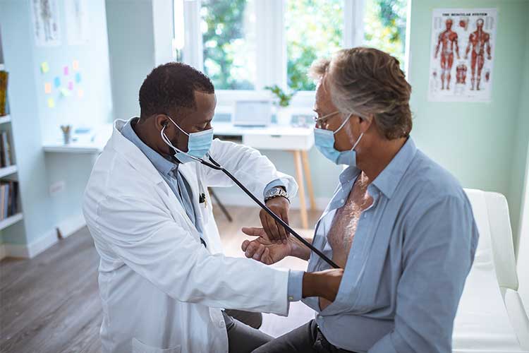 一名黑人男性医生用听诊器听一名白人男性病人的心脏，同时用另一只手测量他的脉搏。两人都戴着口罩。