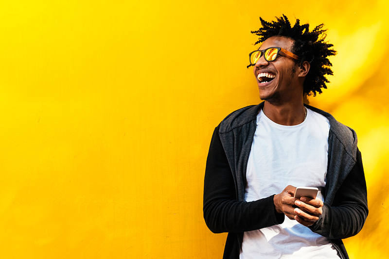 快乐的黑人男子戴着太阳镜在明亮的黄色墙壁前使用手机